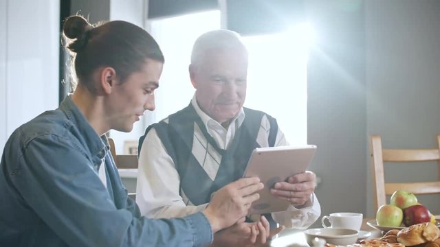 Zoom in shot of happy young man with ponytail showing tablet to grandfather with grey hair and teaching him to use it while having breakfast on sunny morning