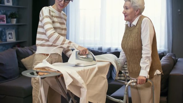 Tilt up of cheerful young woman in glasses ironing shirt on board and chatting with senior lady with walkers