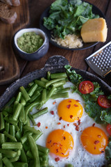 Fried eggs in an old pan with tomatoes, avocado pesto, green beans, pepper and salad on a wooden table, Closeup