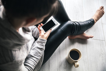 beautiful stylish girl in leather trousers and a modern sweater sitting on the floor with a cup of coffee and mobile phone