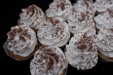 Close-up of many cupcakes on a black background. White cream and chocolate chips. At an angle.