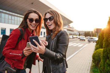 Image of two joyous european women wearing sunglasses looking at smartphone, while standing with...
