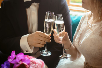 Hands of groom and bride with glasses of champagne at wedding day. Bridal couple hugging. Wedding bouquet at background. Wedding love and family concept close up macro photo with selective focus