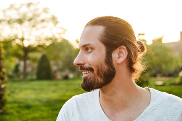 Profile portrait closeup of stylish bearded man 30s with tied hair in white t-shirt smiling, while...