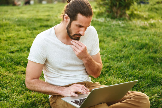 Image of caucasian concentrated man in casual wear sitting on grass in green park and working on silver laptop