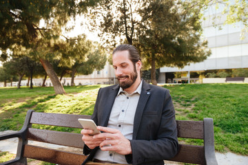 Fototapeta premium Picture of european bearded man 30s in formal wear holding and typing on cell phone, while resting in city park