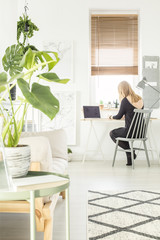Blonde girl sitting by the study corner desk with lamp and laptop in white living room interior with map posters and fresh plants