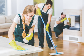 Young member of a cleaning crew wearing green overalls and yellow gloves wiping a white table in apartment interior with the rest of the team in blurred background - obrazy, fototapety, plakaty