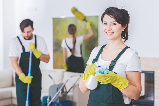 Group of young, hard-working professional cleaners in dirty apartment. Woman with cleaning solution and cloth against blurred background