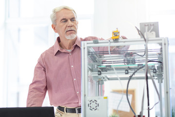 Under supervision. Pleasant senior man working in the office and controlling the performance of a 3D printer