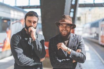 Portrait of two multi-ethnic bearded friends together wearing casual clothes and waiting the train on the railway platform.