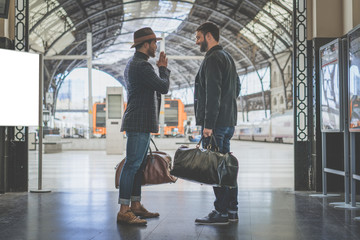 Two confident multi-ethnic bearded businessmans together wearing casual clothes and holding travel bags in hands waiting the train on the railway platform.