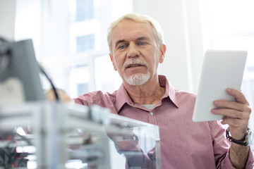 Vital changes. Charming senior office worker changing configurations of 3D printer while reading...