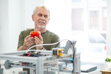 Like the result. Cheerful senior man looking at the printed red tomato printed with 3D printer while being satisfied with the result