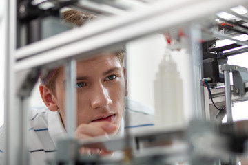 Cutting-edge innovations. Charming young man leaning closely to a 3D printer and watching a little...