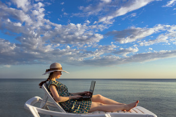 the girl is dressed in a hat and dress, lying on a chaise longue and holding a computer, a freelancer on a blue sea and sky background