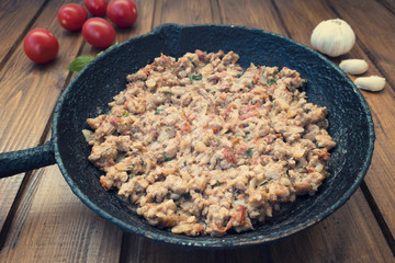 Appetizing fried minced meat with vegetables and greens in rustic old frying pan close-up, tomatoes and chink in the background