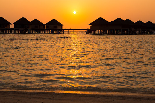 Silhouette of a row of bungalows on a wooden pier at sunset viewed from a sandy beach, Maldives
