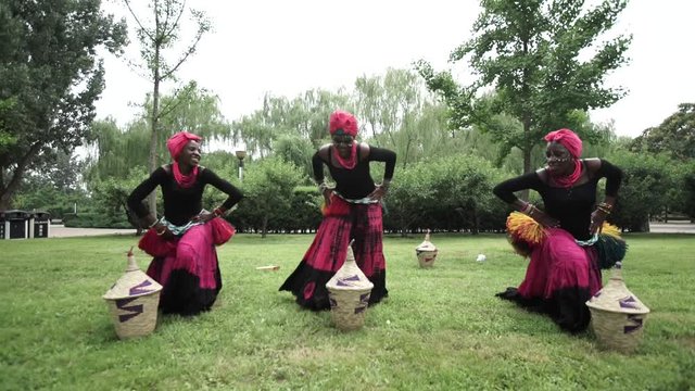 Three african women are dancing a folk traditional dance on a grass meadow, in traditional costumes with wicker baskets, sunny, trees' background, African costumes, traditions, African culture, tribes