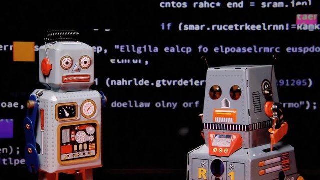 Two tin toy robots standing still in front of a screen showing scrolling source code with a big glitch effect.

