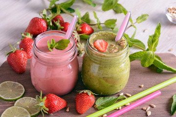 Two jar of bright smoothies surrounded by strawberries and mint stand on a table 