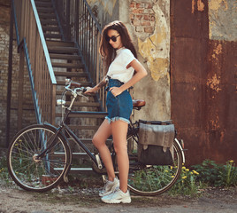 Fototapeta na wymiar Slim girl in short denim shorts, white t-shirt and sunglasses, posing with the city bicycle near an old abandoned brick building.