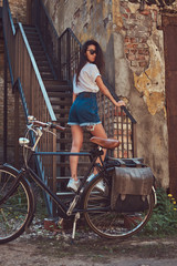Obraz na płótnie Canvas Sexy slim girl wearing short denim shorts and a white t-shirt posing on stairs while standing near the city bicycle against an old abandoned brick building.