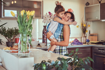 Sweet little girl kisses her mom while sitting in her arms in a kitchen.
