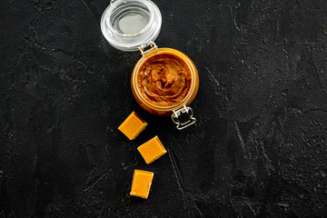 Caramel sauce in glass jar near caramel cubes on black background top view copy space