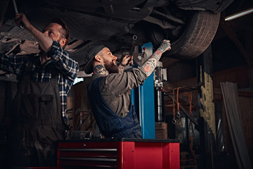 Two bearded auto mechanic in a uniform repair the car's suspension while standing under lifting car in the repair garage. 