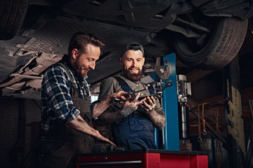 Two auto mechanic in a uniform, working on a workbench while standing under lifting car in the repair garage. 