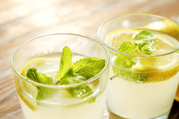 Two glasses full of ice cold refreshing lemonade beverage with mint leaves in sunlight on brown grunged wooden table, slices of ripe organic lemon, whole and halved. Background, copy space, close up.