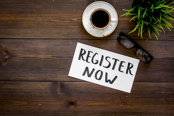 Membership concept. Template for registration. Register now hand lettering iconon word desk with glasses, coffee, plant on dark wooden background top view space for text
