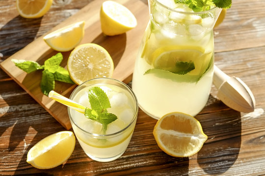 Iced lemonade pitcher, wooden juicer & one glass of cold citrus beverage with lemon slices, mint leaves & yellow straw on brown grunged wood textured table sunny day, background. Close up, copy space.