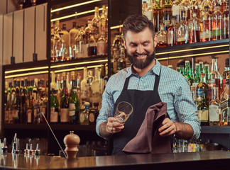 Cheerful stylish brutal barman is cleaning the glass with a cloth at bar counter background.
