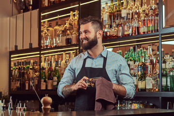 Cheerful stylish brutal barman is cleaning the glass with a cloth at bar counter background.