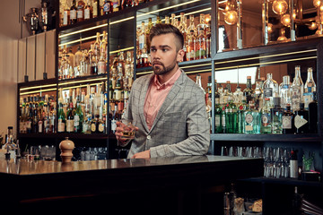 Stylish handsome male in an elegant suit holds a glass of whiskey at bar counter background.