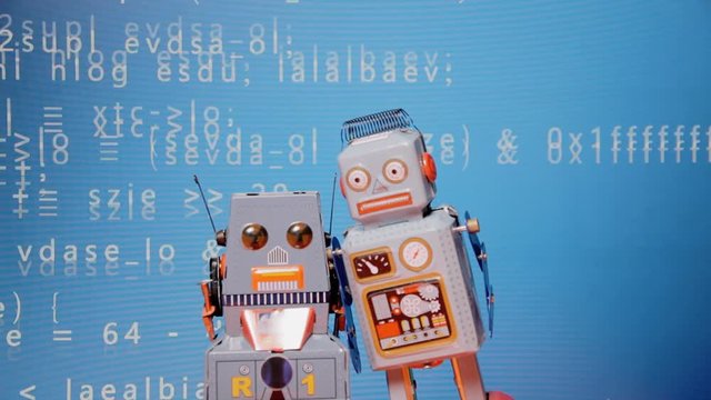 Rack focus shot: two tin toy robots standing still in front of a screen showing scrolling blue source code.
