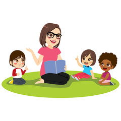 Young female kindergarten teacher explaining lesson reading book to attentive kids sitting on floor