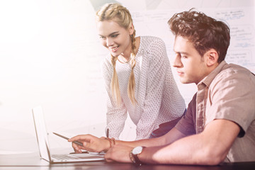 young businessman pointing by pen on laptop screen to smiling businesswoman at modern office