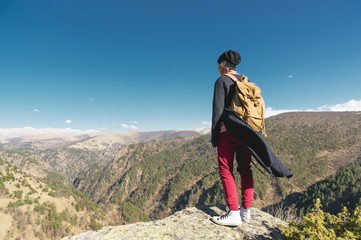Back view of an attractive female tourist, standing in a hat with a backpack on her shoulders enjoying a sunny afternoon in the mountains. The concept of lifestyle travel