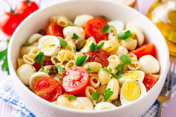 Pasta salad with quail eggs, mozzarella, cherry tomatoes and capers in bowl on purple wooden background