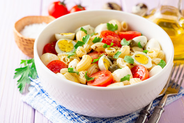 Pasta salad with quail eggs, mozzarella, cherry tomatoes and capers in bowl on purple wooden background