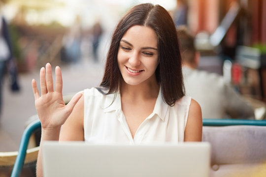 Positive Caucasian female makes video call via laptop computer, waves with hand as sees relatives from abroad, share news, pose against outdoor coffee shop background, uses free interent connection