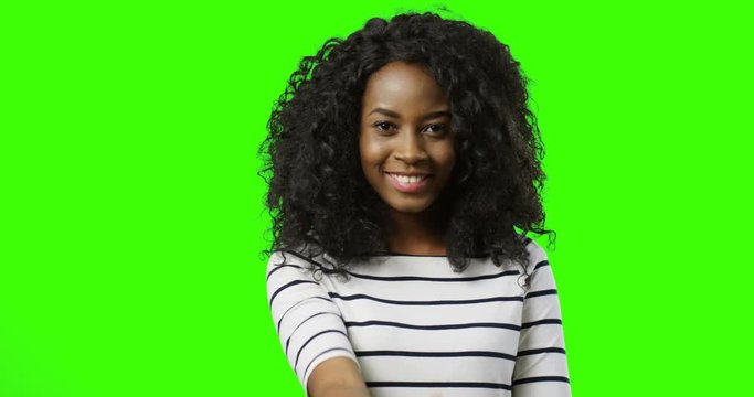 Portrait of the curly African American young woman standing on the green screen background, doing come here gesture with a finger and smiling. Chroma key.
