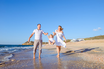 Fototapeta na wymiar Happy family of three - pregnant mother, father and daughter holding hands and having fun walking on the beach. Family vacation, travel concept. Bright sunlight. Copy space.