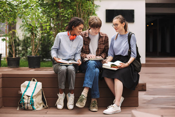 Group of smiling students sitting on bench and reading books in courtyard of university. Young...