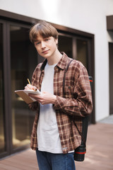 Portrait of young man standing with notebook and pencil in hands and dreamily looking in camera in courtyard of university