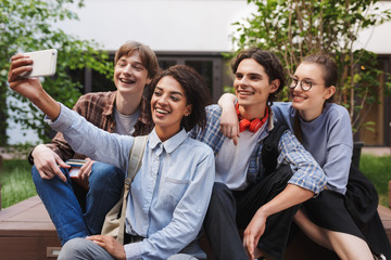 Group of cheerful students sitting and taking cute photos on cellphone while spending time together in courtyard of university
