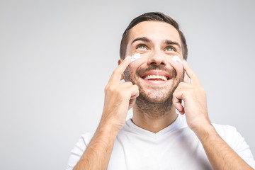 Skin care. Handsome young shirtless man applying cream at his face and looking at himself with smile while standing over gray background and looking up. Close-Up. Space for text.